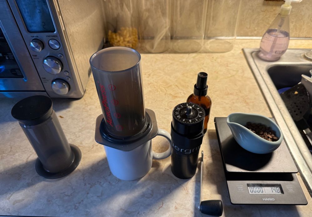 A photo of a coffee mug with an Aeropress, scale weighing coffee beans for one cup, hand coffee grinder, and water spritzer (for the beans).