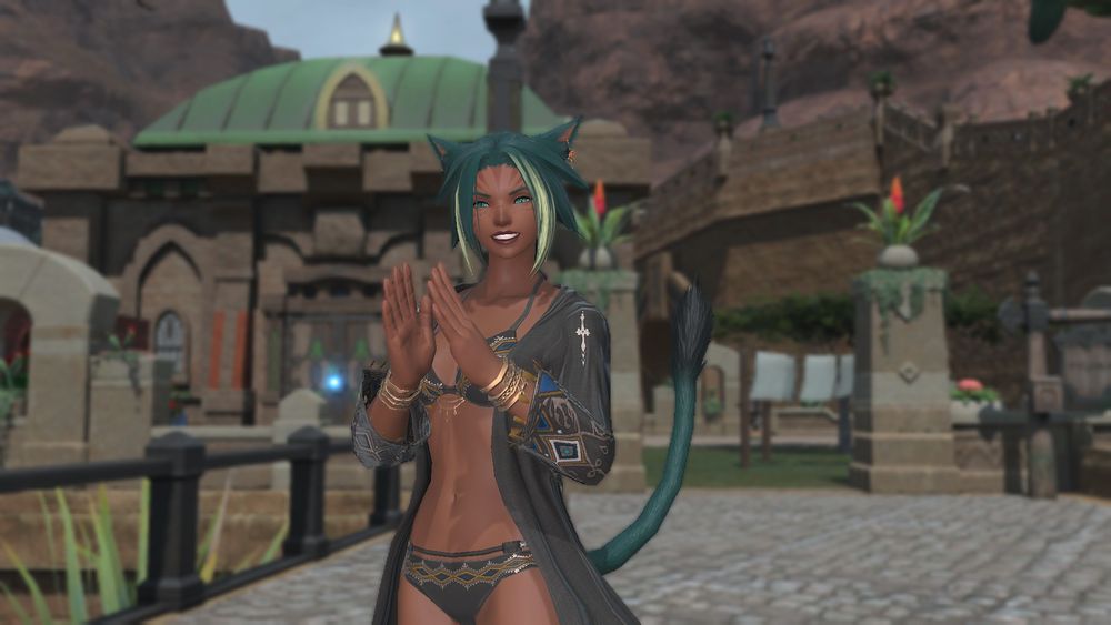 A female miqo'te from the game Final Fantasy 14.  She has medium brown skin, green hair, and green fur on her tails and ears.  She is wearing a black bikini and a cover up top.  She's smiling in front of a house in a dessert area.  