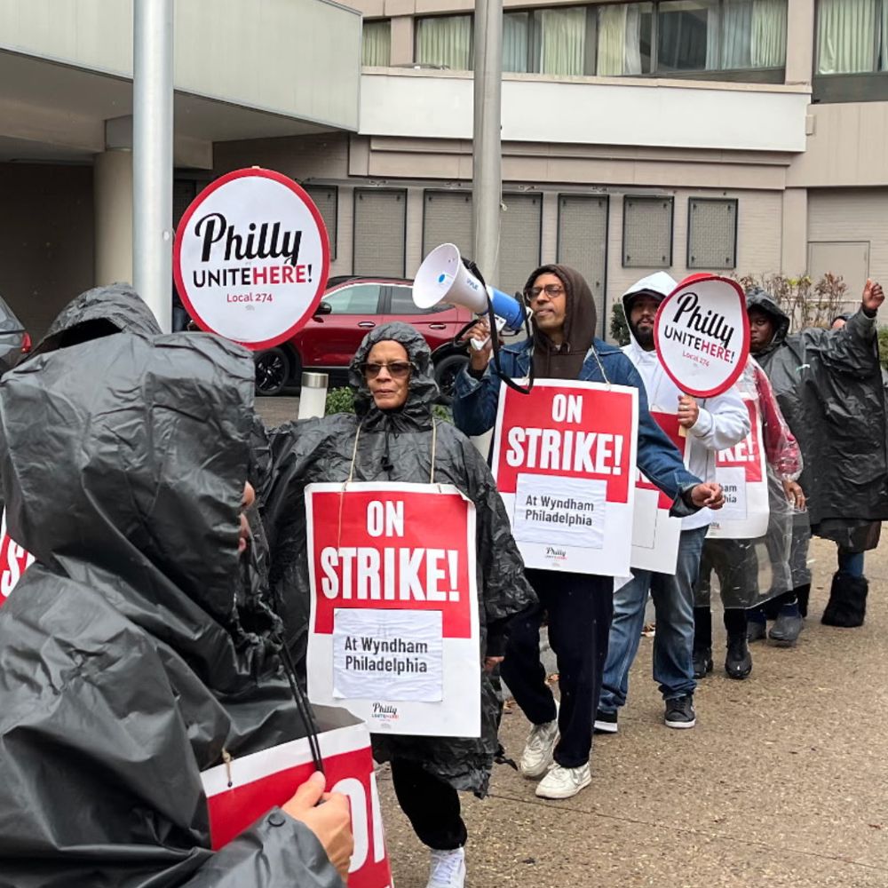 Senator Saval joins striking workers with UNITEHERE Local 274 on the picket line outside the Wyndham Hotel. Workers are wearing raincoats and ponchos to protect against the weather and holding red and white signs that say "ON STRIKE!" Senator Saval is holding a bullhorn and wearing a hoodie to keep off the rain.