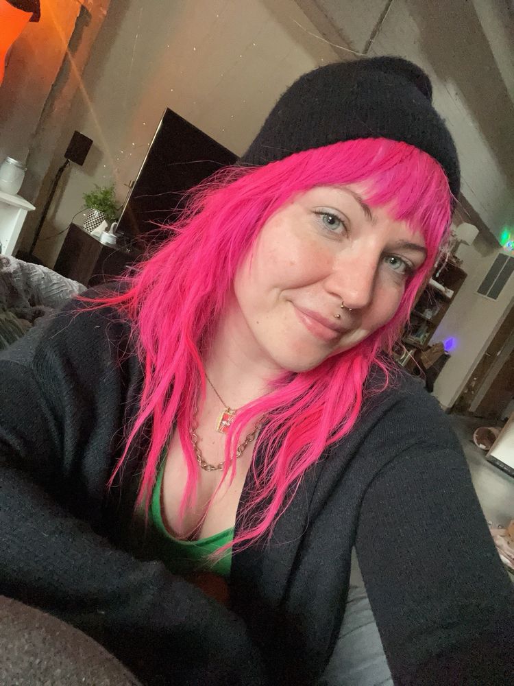 girl looking at the camera with a head tilt & smirk. she has long pink hair, with a black beanie on top of her head. there are two necklaces around her neck. she’s wearing a grey long sleeved sweater over a green tank top 
in the background is random clutter from the living room. 