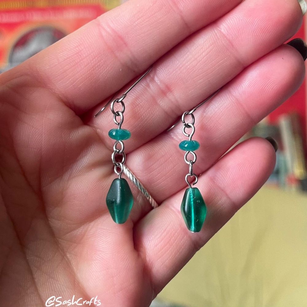 pair of double drop dangly earrings held up between fingers. there are two beads per earring, starting with a small rondelle, flower detail green bead which connects to a larger forest green diamond shape bead. made using glass, titanium, and stainless steel materials. skin friendly!