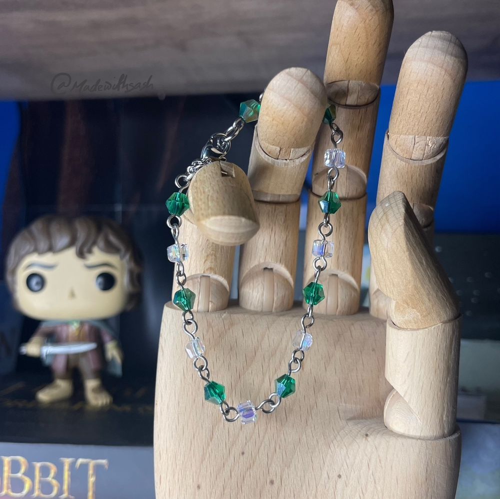 a green and clear/white beaded chain bracelet hanging off a wooden hand mannequin with tolkien books blurred in the background. this is made using glass 6mm forest green bicone beads and 4mm clear cube beads. the beads are singular and alternat between the two, connected with titanium wire loops. the closing is stainless steel jump rings, extension chain and a lobster clasp. sensitive skin friendly!