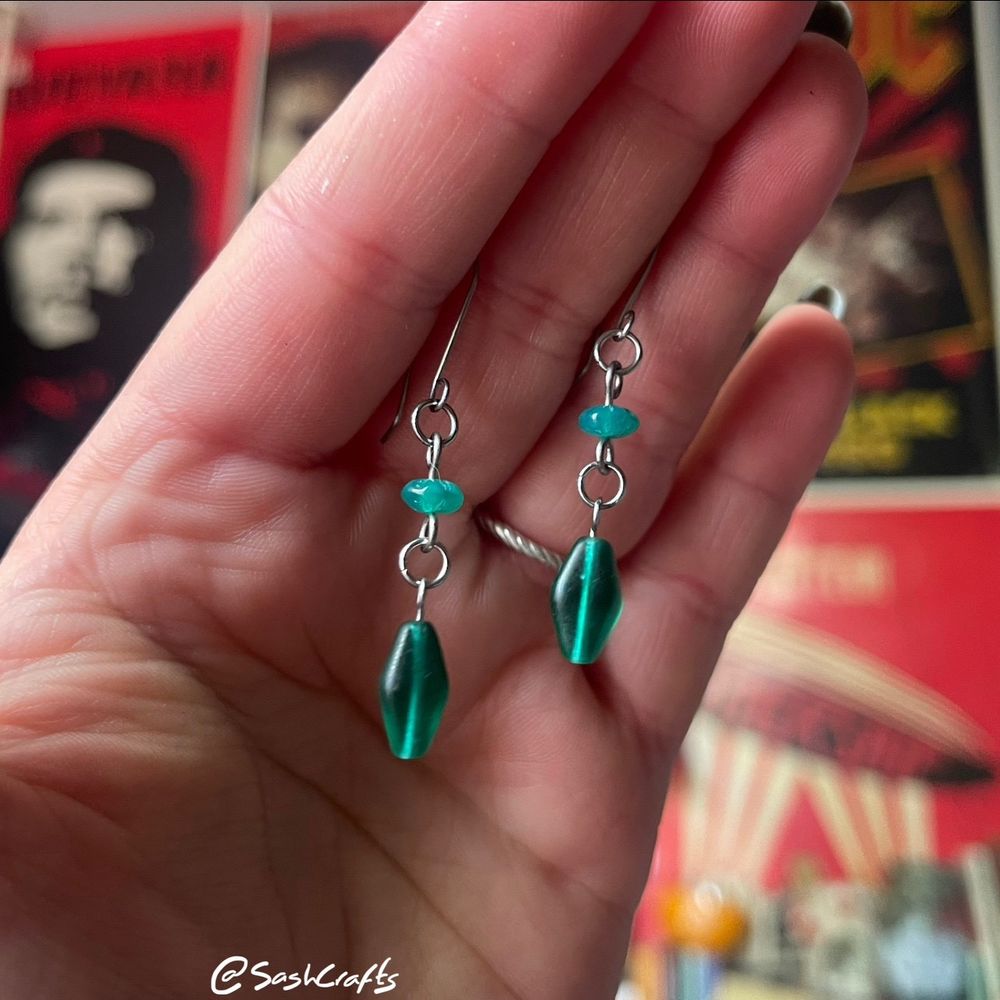 pair of green drop dangly earrings held up between fingers. there are two beads per earring, starting with a small rondelle, flower detail green bead which connects to a larger forest green diamond shape bead. they are a gorgeous emerald green colour. made using glass, titanium, and stainless steel materials. skin friendly!