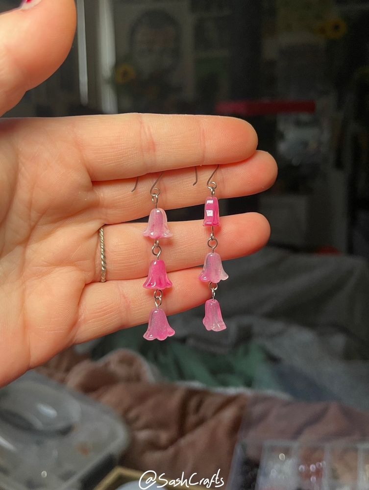 a left pale hand holding up a pair of pink dangly earrings between the first two fingers. same as the previous, the earrings each hold a string of three bell shape flower glass beads that are almost translucent and a beautiful mix of pinks. they’re so pretty!!