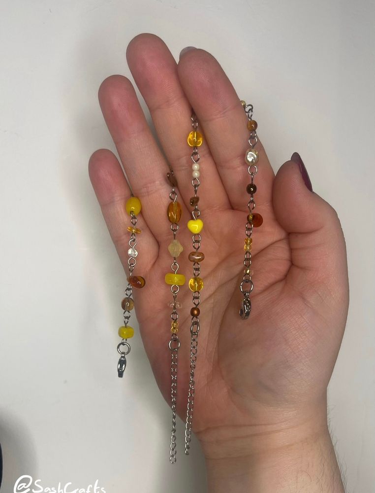 pale right hand holding up two unclasped chain bracelet with yellow beads hanging over the forefinger and the ring finger, palm facing us. the bracelets dangles vertically down either side. the bracelets do not match in order but the beads are from the same selection and variety of yellow and brown. stunning