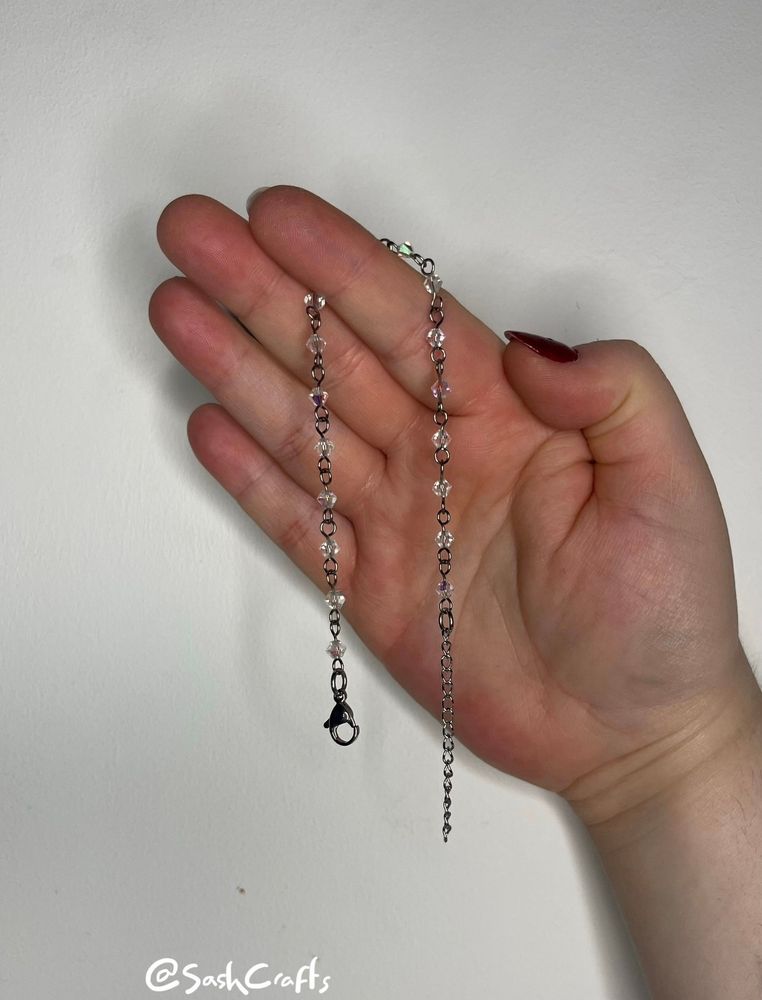 a pale right hand with red nails holding up a bracelet which is unclasped and dangling down either side the forefinger. the bracelet consists of clear suncatcher beads. this bracelet is made using glass beads, titanium wire, and stainless steel closures and extension. 