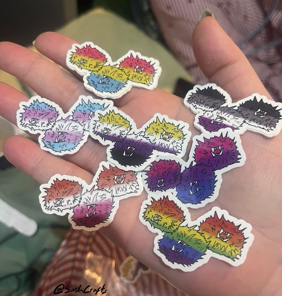 a pale hand holding out each of the seven pride tribbles stickers. the design is the same but the flags are not, from top to bottom is pansexual, asexual, trans, nonbinary, bisexual, lesbian and the 6 colour pride flag