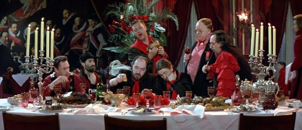 Men in red and black at an ornate dining table. They look the confusing of worthless rich people at the bounty before them, too stupidly drunk from their stolen wealth to appreciate the value abstracted in the food before them. The still is from the film THE COOK, THE THIEF, HIS WIFE, AND HER LOVER.