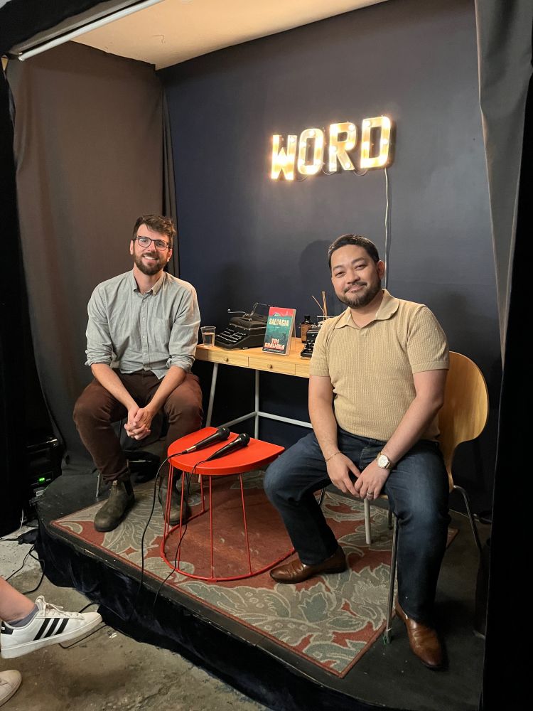 Extremely cool JAB clients Tim Chawaga (left) and Victor Manibo (right) sitting on the stage at WORD Bookstore and smiling for the camera. A copy of SALVAGIA by Tim Chawaga is displayed between them, and a lighted sign of the bookstore's name, WORD, hangs on the wall behind them.