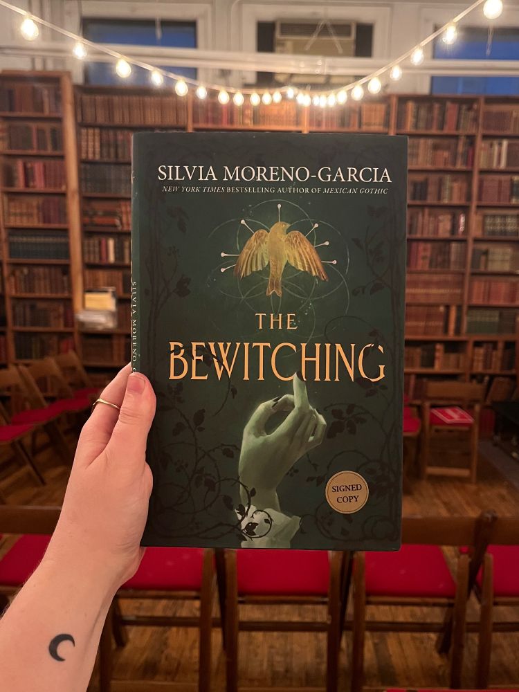A hand with a small moon tattoo holding up THE BEWITCHING by Silvia Moreno-Garcia against a backdrop of chairs from the signing and bookshelves at The Strand