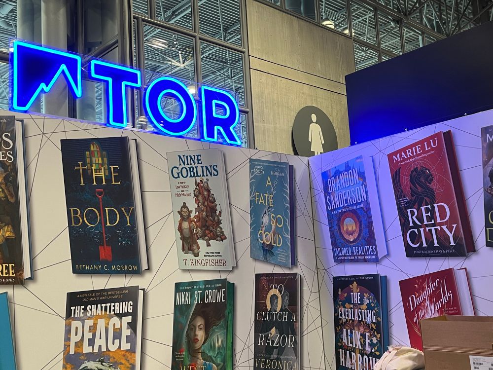 The Tor section at the Macmillan booth. A blue neon sign reads Tor on the top of the booth. Printed on the wall are various Tor titles, including TAILORED REALITIES by JAB author Brandon Sanderson, forthcoming this December