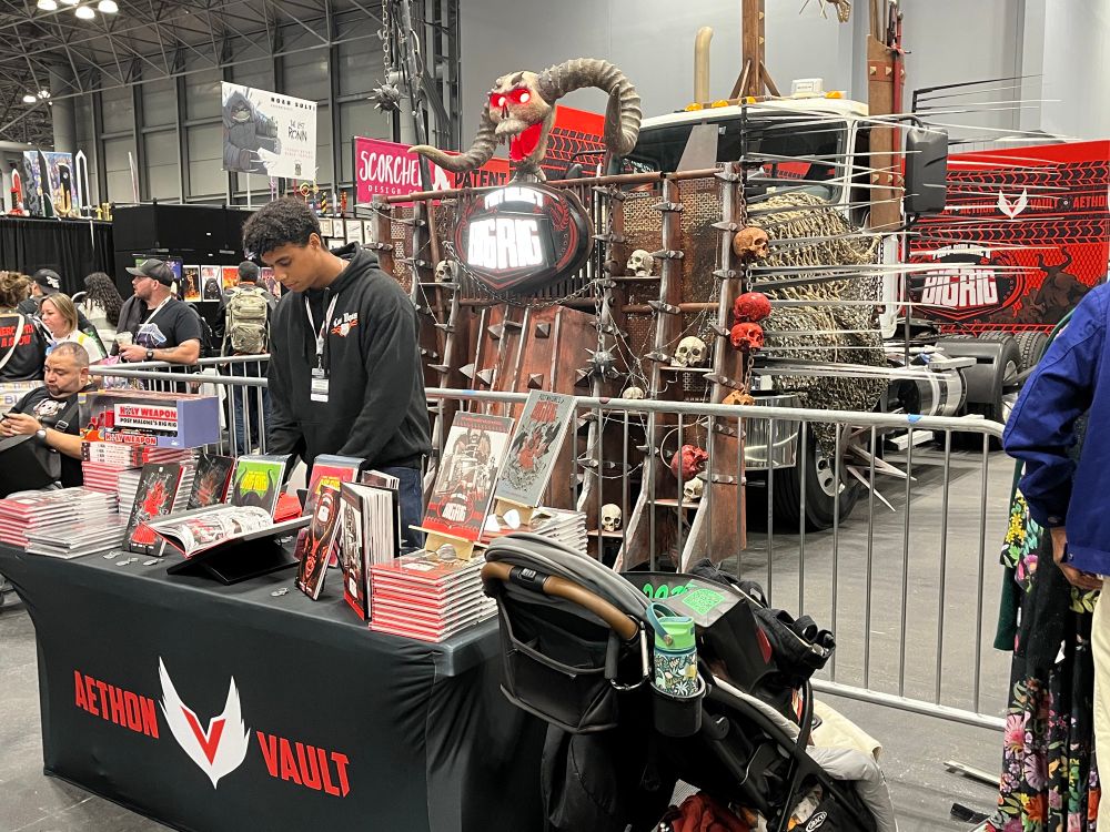 In the background, Post Malone’s BIGRIG but made reality: a giant, decked-out truck with skulls and spikes and seriously cool features (oh my!). In the foreground, a small table featuring the BIGRIG comic and Aethon and Vault’s names