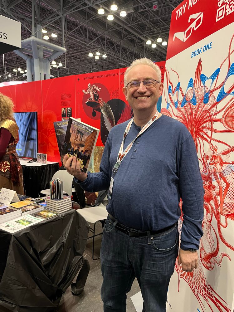 JAB author Alex Shvartsman standing in front of booth 3325 and holding up copies of his books, THE MIDDLING AFFLICTION and KAKISTOCRACY