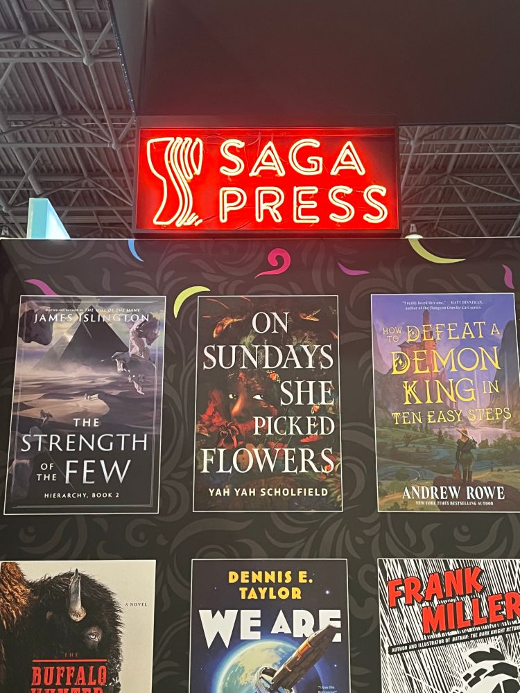 The Saga Press section at the Simon & Schuster booth. A red neon sign reads Saga Press on the top of the booth. Printed on the wall are various Saga titles, including ON SUNDAYS SHE PICKED FLOWERS, the debut horror from JAB client Yah Yah Scholfield