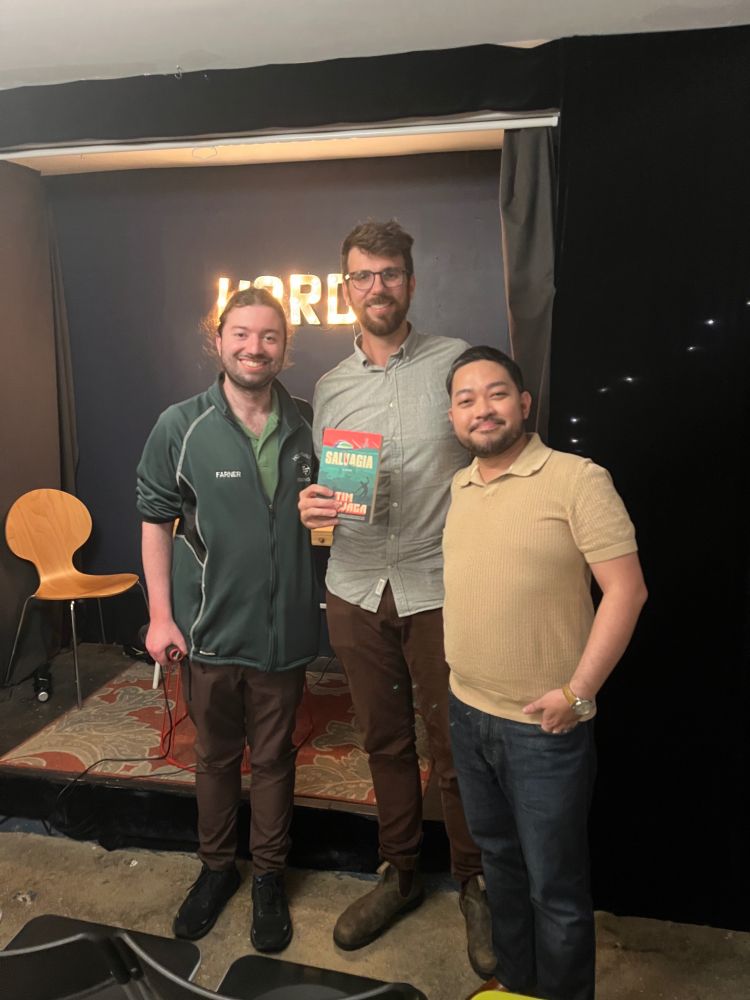 Extremely cool JAB agent James Farner (left), and JAB clients Tim Chawaga (center) and Victor Manibo (right) posing in front of the stage at WORD Bookstore. Tim is holding a copy of his debut SALVAGIA by Tim Chawaga, and a lighted sign of the bookstore's name, WORD, hangs on the wall behind them.