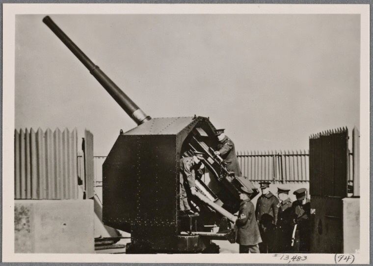 Black and white photo of a large anti-aircraft cannon in peacetime with six uniformed men. It appears to be armored on all but the back side.