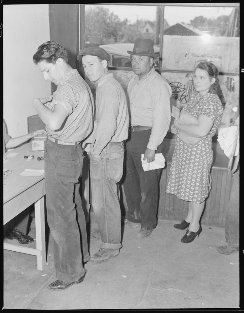 Black and white photograph of three men and one woman at the front of a food bank line. The man at the very front is looking in his pocket. One arm and one shoe of a man behind the desk is visible, also a small box of some kind and some forms and ink stamps.