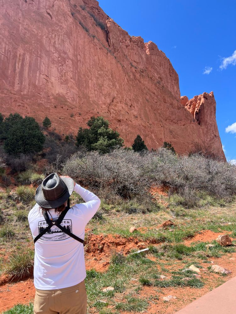 Birder looks up at a high red cliff with binoculars against a blue sky