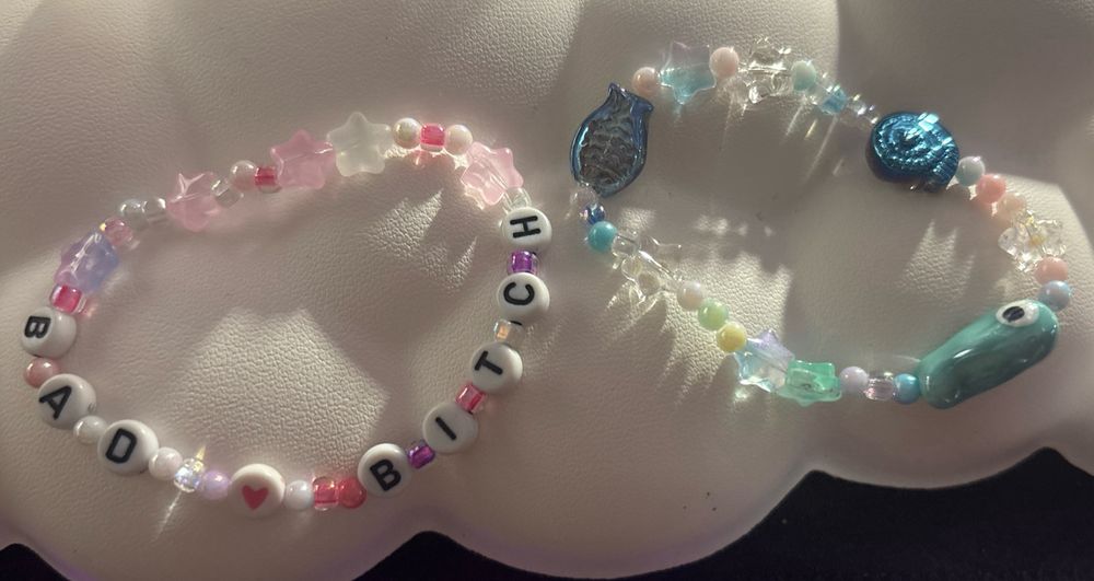 Two beaded bracelets. The one on the left is pink with beads that read out “bad bitch”. The right one is blue and white with fish beads. 