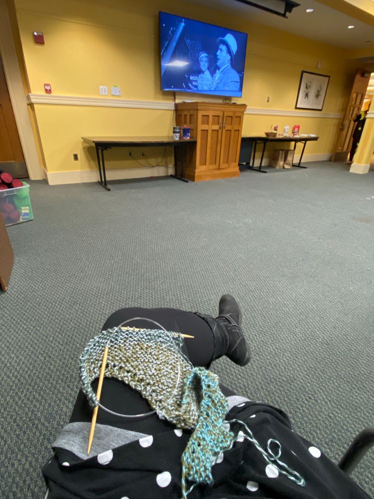 A view of a woman’s legs crossed with a partially knitted scarf on her lap. In the background is a projector with a movie playing 
