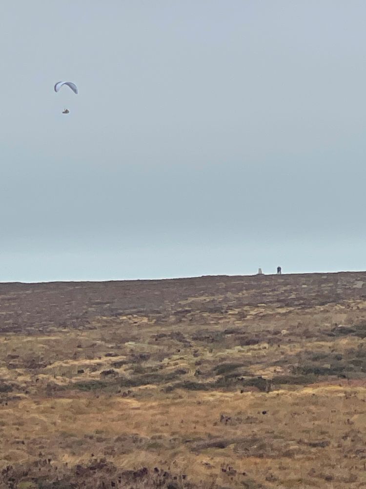 Unexpected sight: Parasailers at the far northern tip of the ridge, above Hay-on-Wye. Brown, barren ridge top with cloudy sky in the background. Tiny figures on the horizon, and a lone flyer in the sky. 