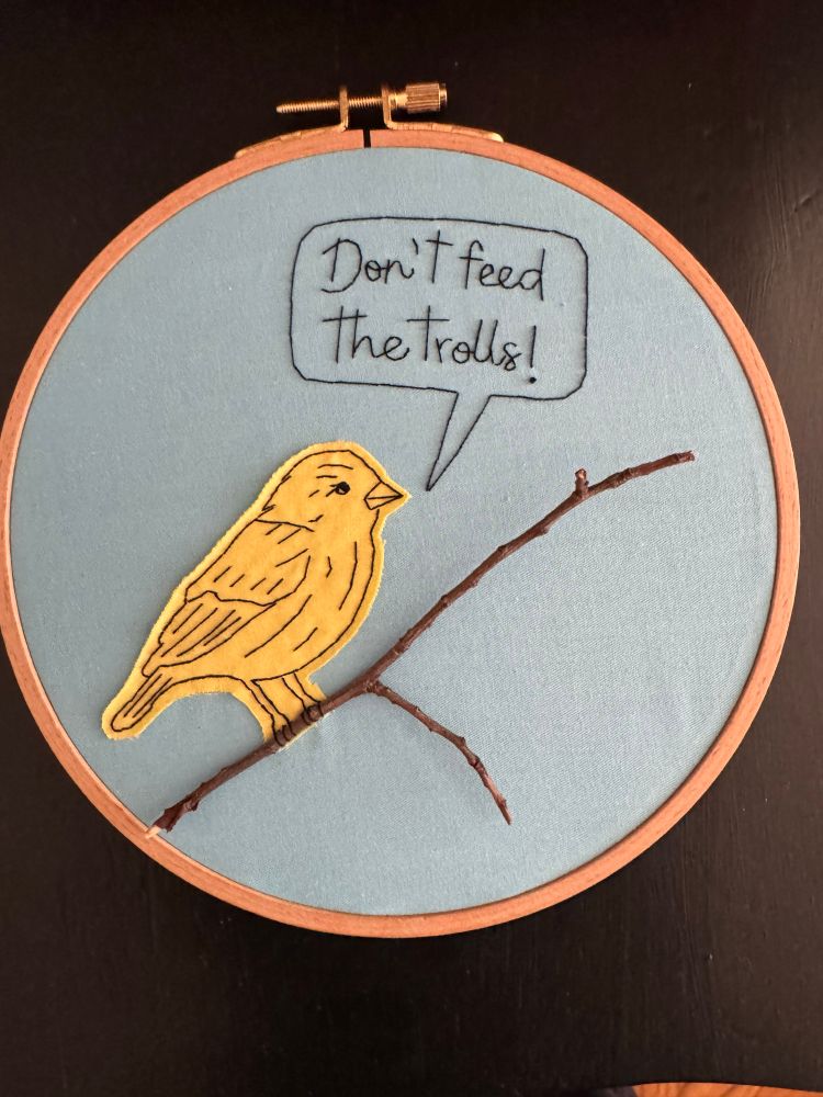 An embroidery hoop with An appliquéd yellow cardinal sitting on a branch made from an actual Stick with a quote bubble that reads: don’t feed the trolls 