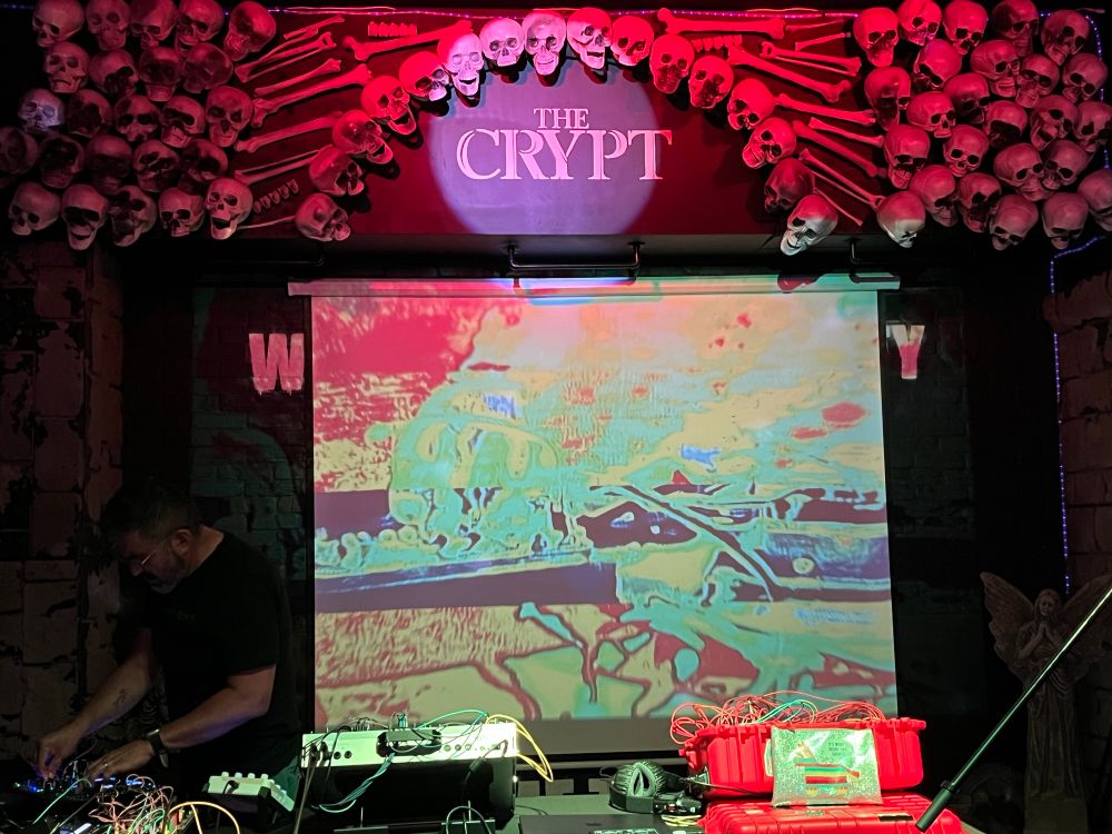 Sine Mountain at his modular synth, in shadow far left with abstract color screen centered under bone-intensive venue logo above the stage.
