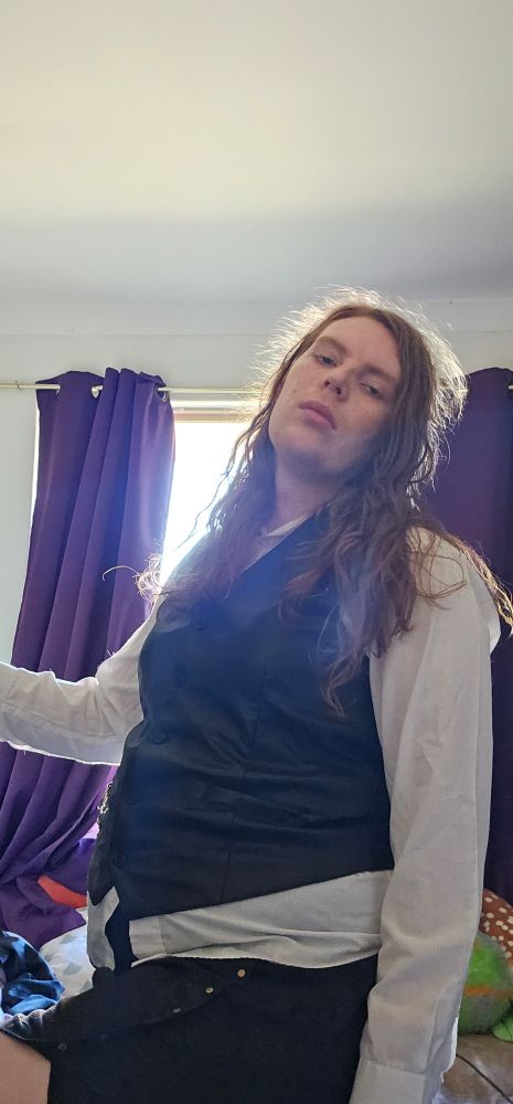 Vivian wearing a black leather vest (without a chain) on top of a longsleeve white button up. Xe is also wearing A short black skirt that only  goes halfway down her thighs