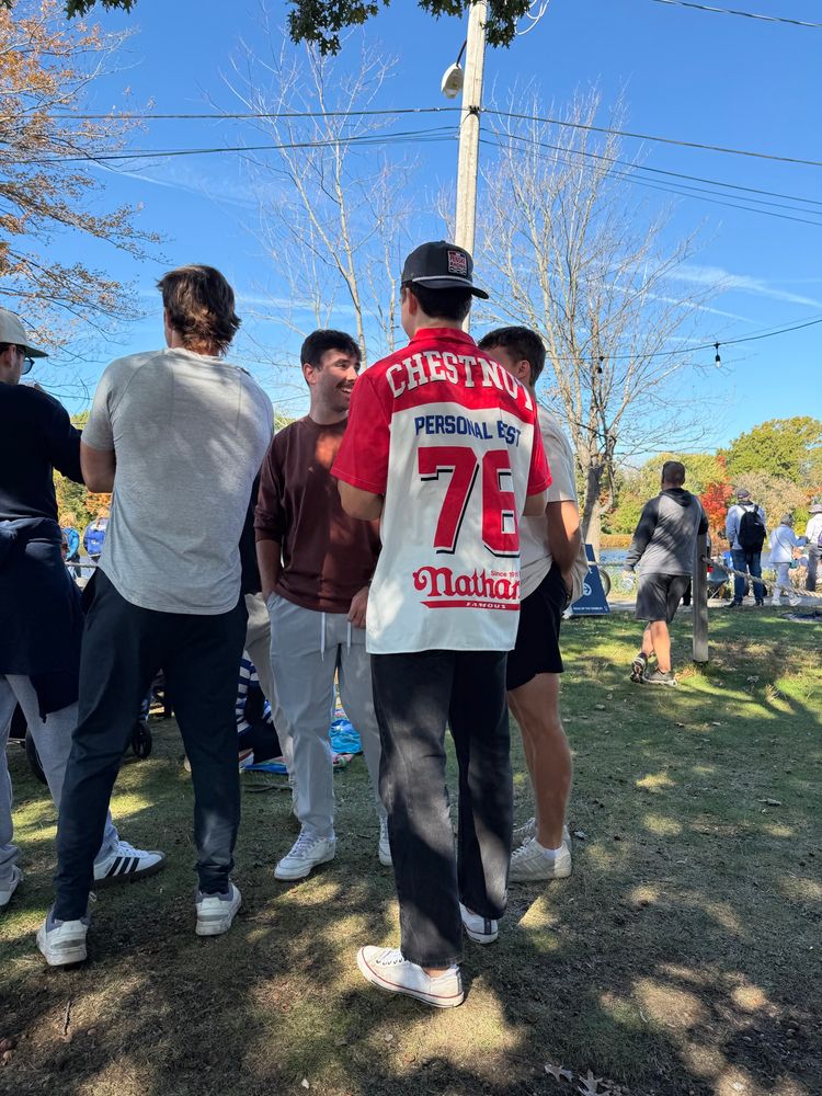 Boston College alumni standing in a circle planning to watch the UConn game and being egged on by their ringleader who, for unexplained reasons, is wearing a Joey Chestnutt jersey celebrating Joey’s personal record of 76 hot dogs eaten at the Nathan’s Famous Hot Dog Eating Contest