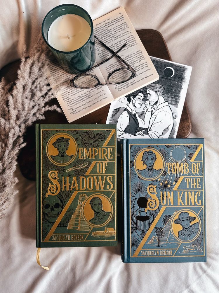 Photograph of vintage clothbound gold foiled editions of Empire of Shadows and Tomb of the Sun King on cozy bed with art print, candle, flowers, and glasses