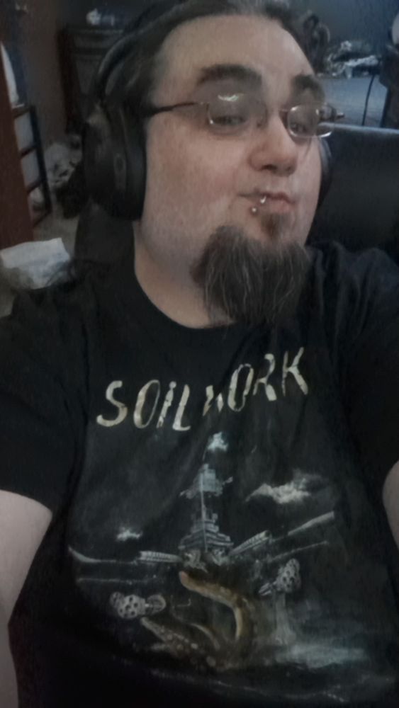 A man with long black hair pulled back in a ponytail and a goatee, wearing glasses, headphones and the t-shirt mentioned in the post. The shirt features the name of the band soilwork and depicts a scene of a naval ship at sea with dark skies and a kraken with its tentacles wrapped around the bow of the ship. The kraken's head is under the water and not visible. 