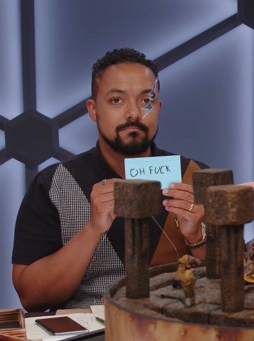 an image of Jasper William Cartwright, a black man with short curly hair and a blue lightning bolt painted over his left eye, staring into the camera with a neutral expression while holding up a small note card that says "oh fuck"