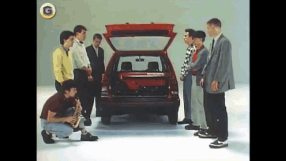 a group of men are standing around a red car with the trunk open and a g on the bottom right