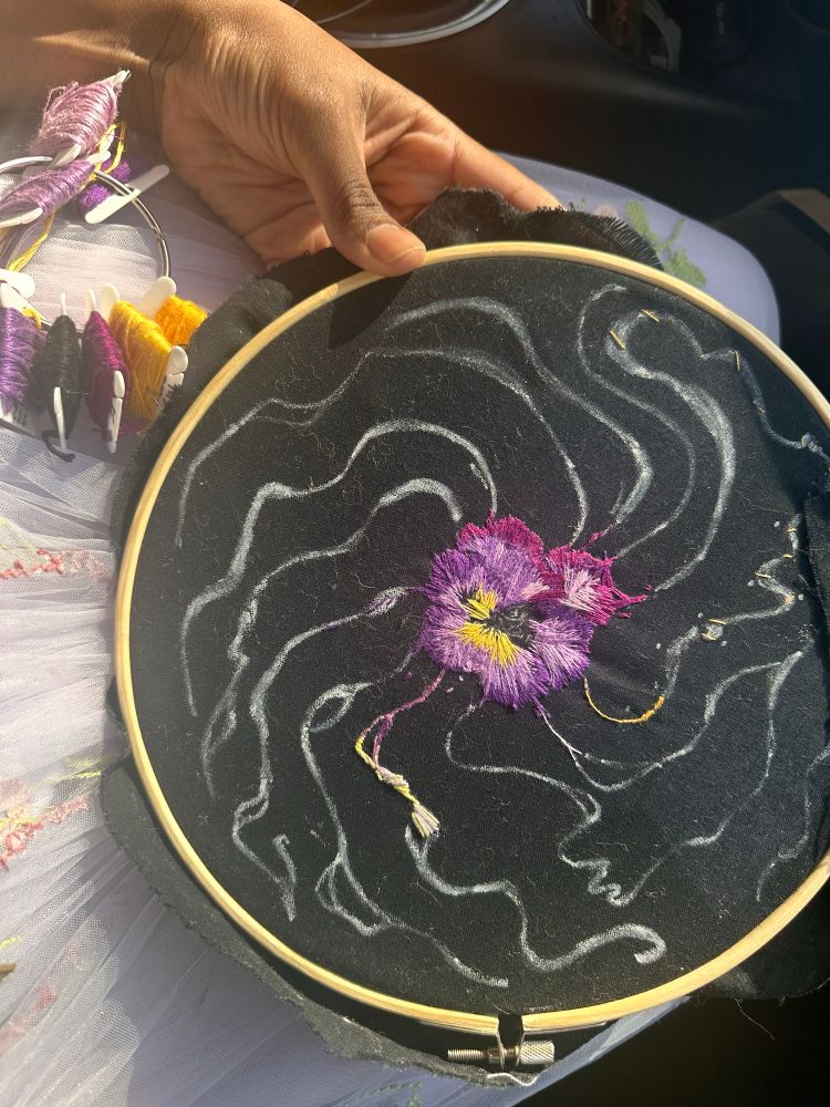More progress was made on the embroidered pansy. Embroidery thread on top of a purple floral skirt next to the embroidery hoop work in progress pansy. 