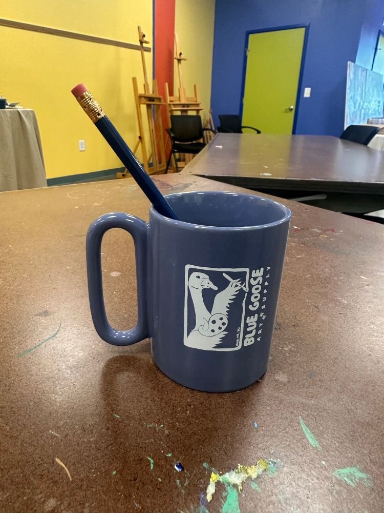 A blue mug with the Blue Goose Art Supply logo sits on a table with a pencil in it