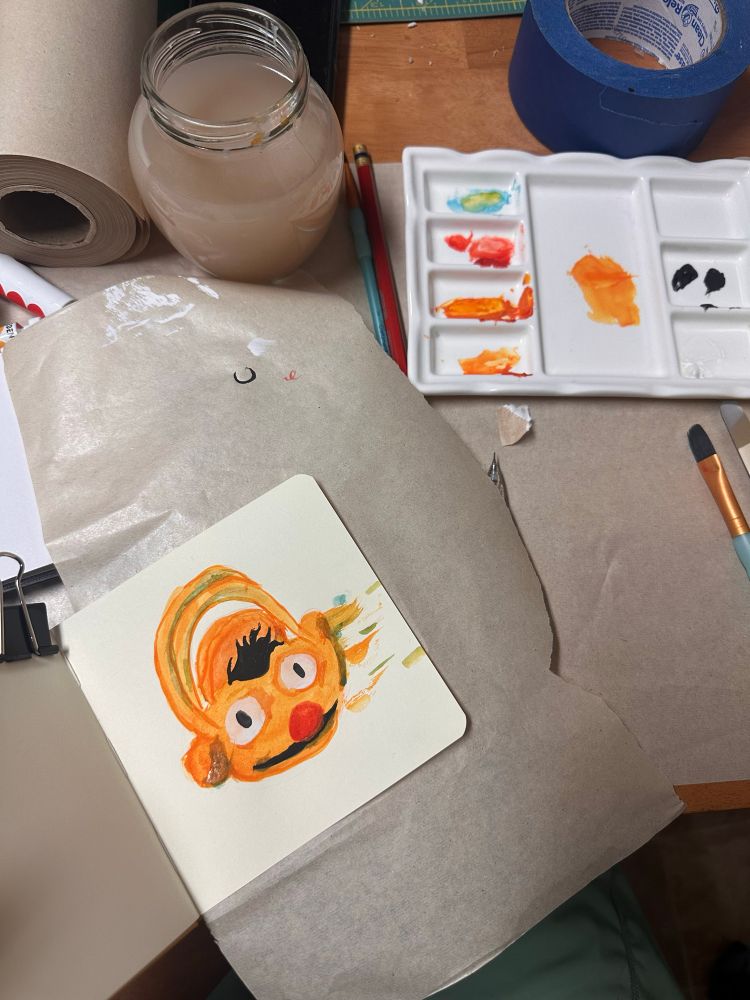 Gouache paint, a ceramic played and a sketchbook are scattered in a table. The sketchbook has a fresh painting of a Bert from Sesame Street candy basket