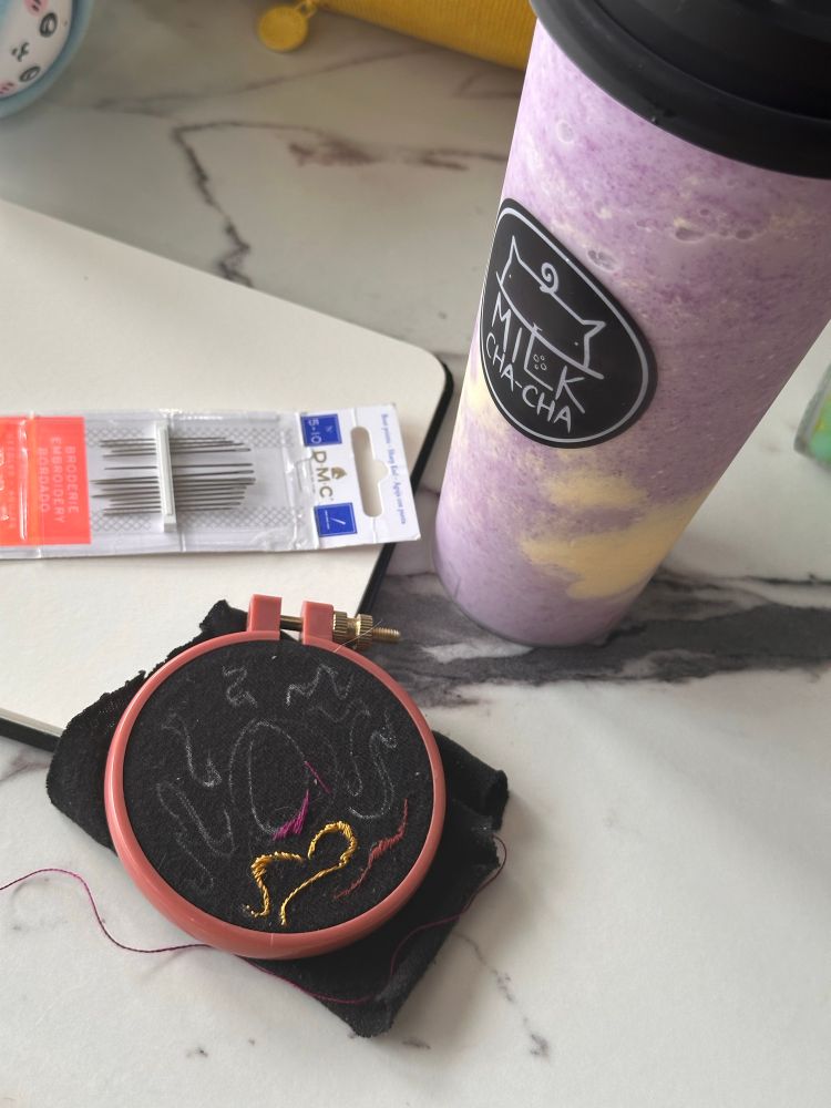 An embroidery hoop and a cup of ube slush sit next to embroidery needles and a sketchbook on a marble table 