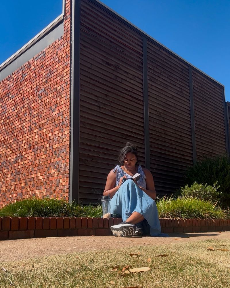 A Black woman sits on a brick step in front of a business. She is drawing on a small sketchbook with a homemade iced coffee next to her.