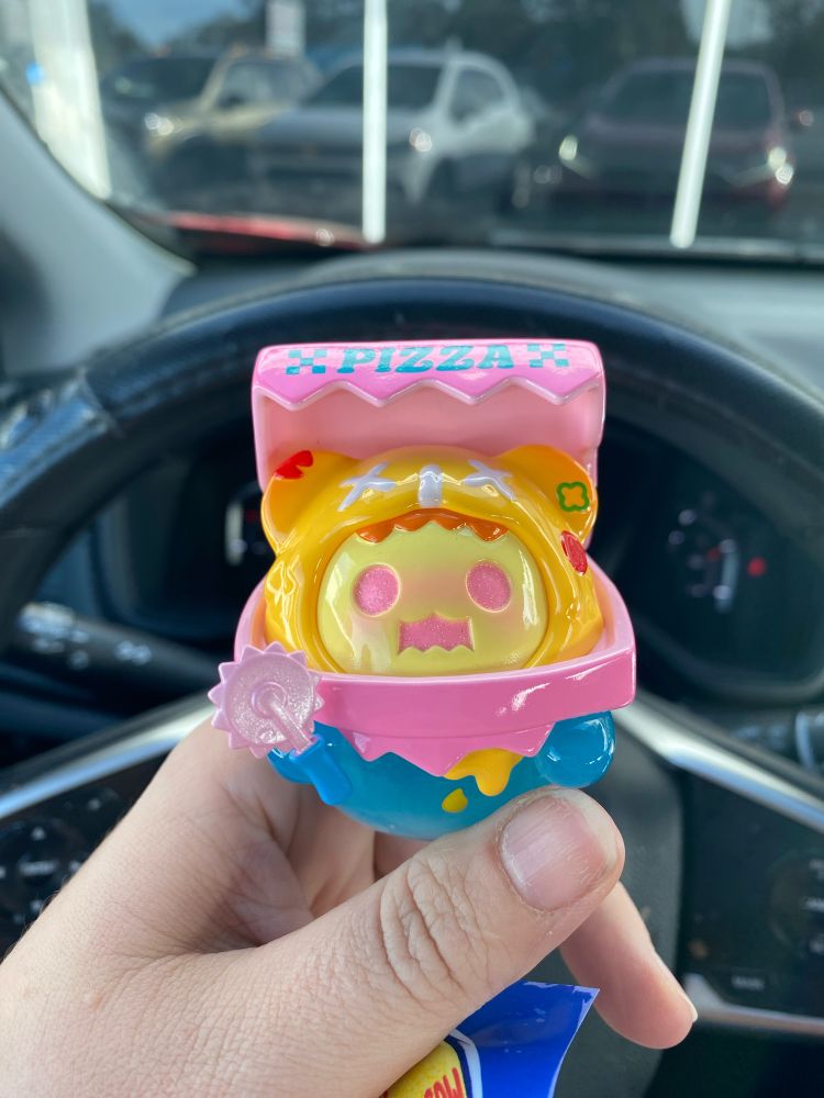 photo of a figure from shinwoo's baby ghost bear line, the diner series; figure is a small semi-translucent blue ghost with a pale yellow face and pink glitter eyes. it holds a pizza cutter and has a pizza box eating its head.