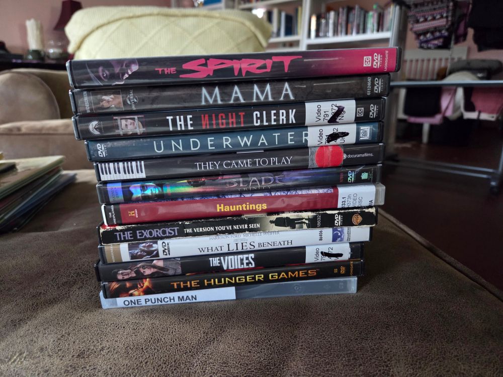 A stack of DVDs on a couch cushion. They are The Spirit, Guillermo Del Toro's Mama, The Night Clerk, Underwater, They Came To Play, Blade Trinity, the History Channel's Hauntings, The Exorcist, What Lies Beneath, The Voices, The Hunger Games, and One Punch Man in a case that is clearly not the original.
