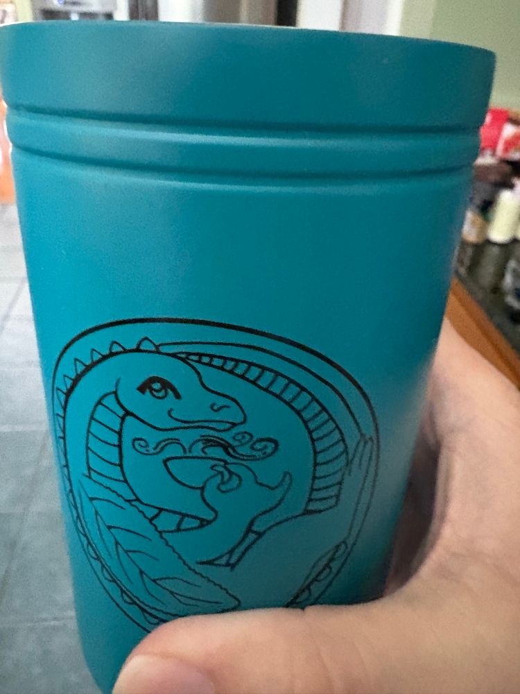 A white woman’s hand holding a green travel mug with a tea leaf-winged dragon on it outlined in black. The dragon is holding a steaming mug of tea.