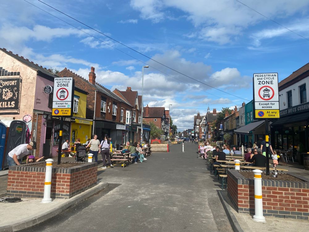 Brick planters, bollards and signage with text and symbols (pedestrian and cycle zone - no motorbike or vehicles at any time except for access. No parking at any time) at the entrance to a street full of people. 