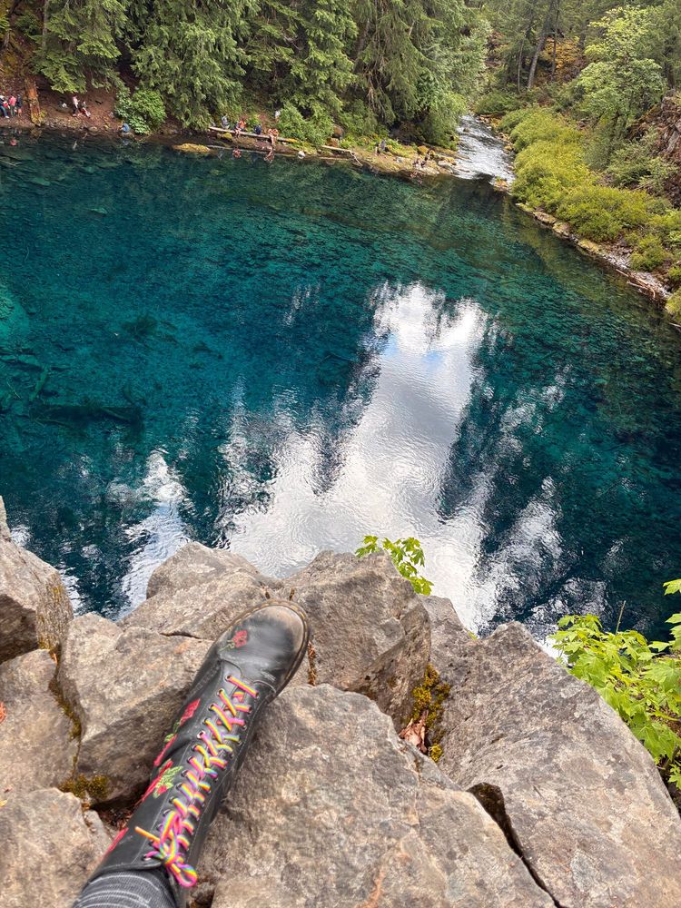an overhead shot of the deep clear blue pool at Tamolitch Oregon, with clouds and trees reflected in it. also my left boot with the rainbow laces is in the shot