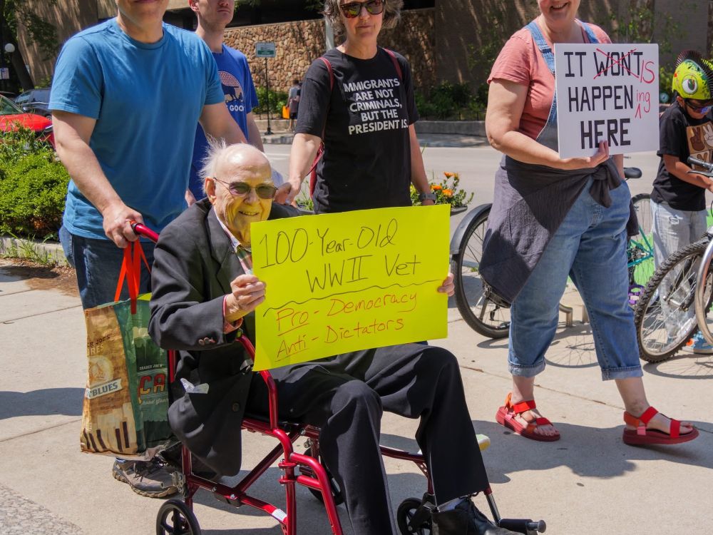 An elderly man in a suit and sunglasses, in a red wheelchair, at a protest in oak Park, Illinois. The man is holding a sign that says “100 year old WWII vet, pro-democracy anti-dictators”. 