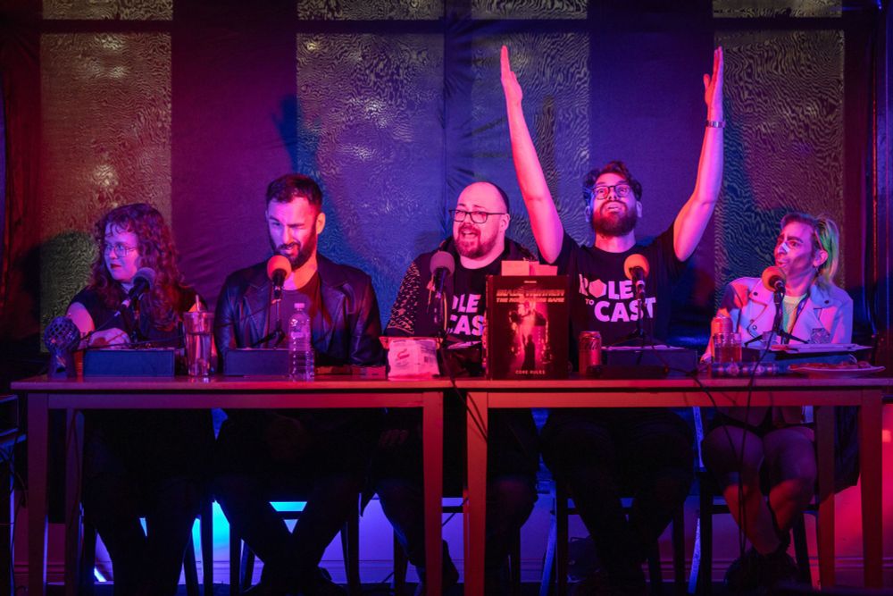 From left to right:
Zola, Sean, Phil, Chris and Ellen pose at their table for the Role to Cast live show
In front of them are microphones and the Blade Runner TTRPG book
