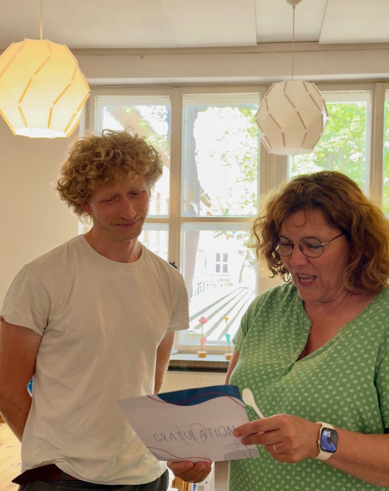 Das Bild zeigt eine erwachsene männliche Person mit blonden Locken in weißem T-Shirt, die auf ein Blatt Papier mit der Aufschrift "Gratulation 10" schaut, welches von einer erwachsenen weiblichen Person mit Locken und Brille in grünem Oberteil gehalten wird. Über den beiden hängen Lampen, von denen eine leuchtet und sie stehen vor einem Fenster.