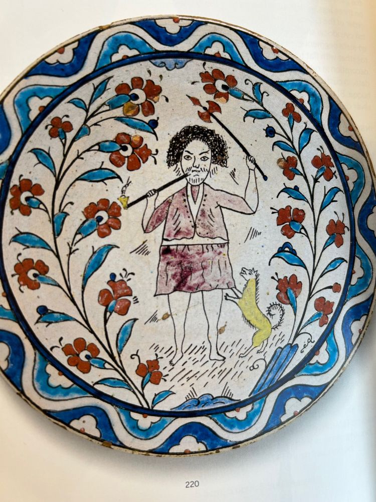 Iznik pottery plate featuring blue patterned borders, blue vines with red flowers, and a scraggly-looking figure in the middle, holding an axe in one hand and a long pipe in the other. A yellow dog is trying to climb his leg.