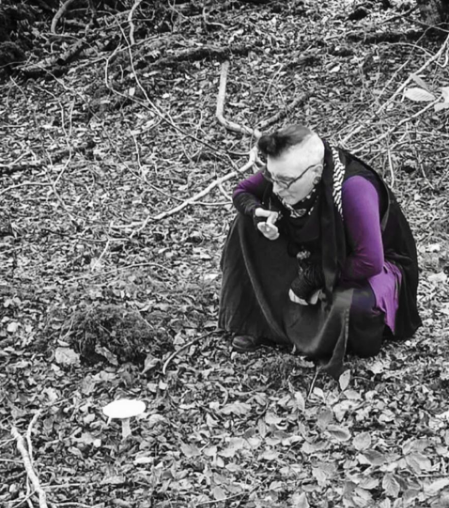a queer person kneeling in the woods is gazing upon a single white mushroom. the image is black & white except the person's purple clothes. 