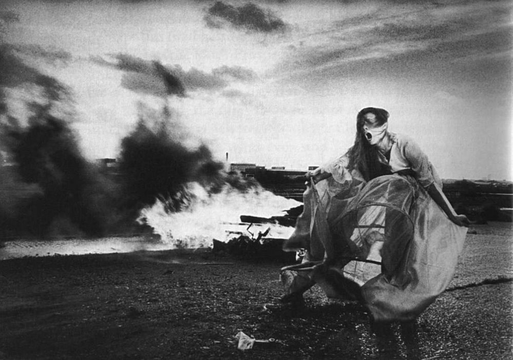 Black and white film still of an open sky and stark landscape. a human figure in a billowing sheer gown screams with their full body in front of a large fire. The image is Tilda Swinton in The Last of England by Derek Jarman 1987