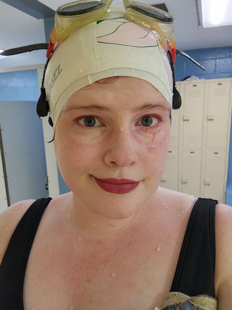 A woman wearing a swimming, she has marks from goggles around her eyes and red makeup dripping slightly under one eye